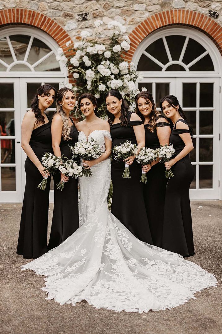 Bride in off the shoulder lace wedding dress with puddle train standing with bridesmaids in black strappy bridesmaid dresses 