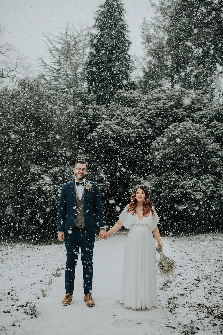 Groom wearing navy suit with patterned waistcoat with bride in a short sleeve wedding dress with sparkly belt detail at snowy winter wedding 