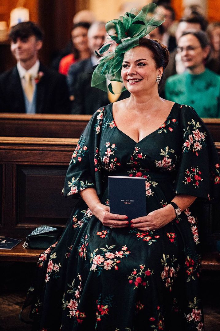 Wedding guest sitting on a church pew wearing a green wedding fascinator and floral dress 