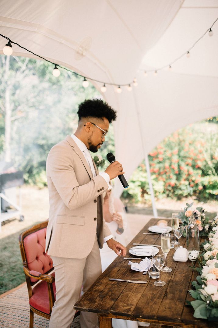 Groom in cream suit giving a speech at outdoor BBQ wedding