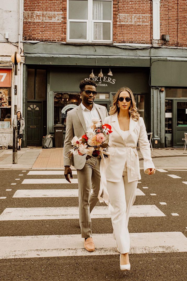 bride in white wedding jumpsuit walks across the road with her groom both wearing sunglasses 