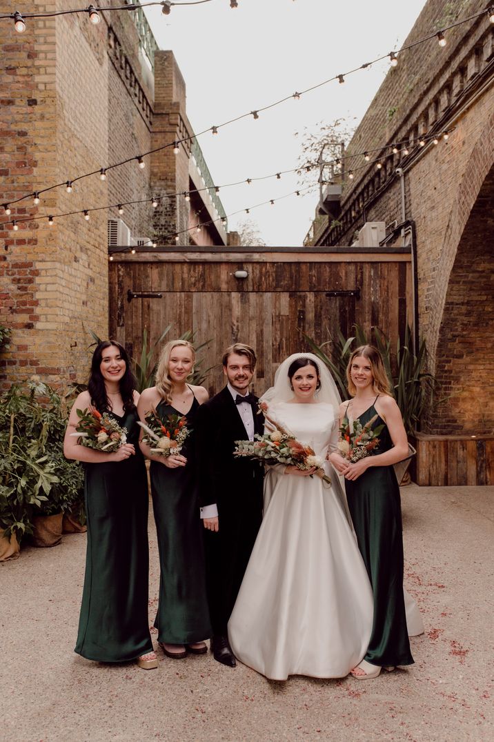 Bride and groom standing with bridal party wearing bottle green satin strappy bridesmaid dresses from Rewritten Bridesmaids, bride to be gift box 