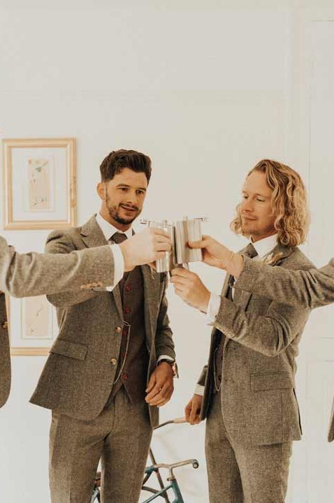 Groom and groomsmen in matching marl grey three piece suits cheersing their hip flasks at Anran Devon Wedding 