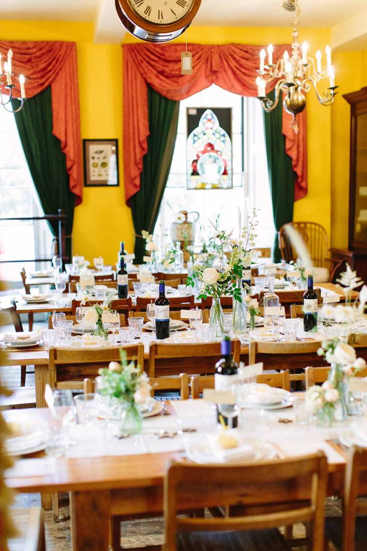 Classic rustic wedding tablescape with dried flower and white rose centrepieces, white table runners, red and green curtains and large clock with gold chandeliers at Brunswick House London wedding venue 