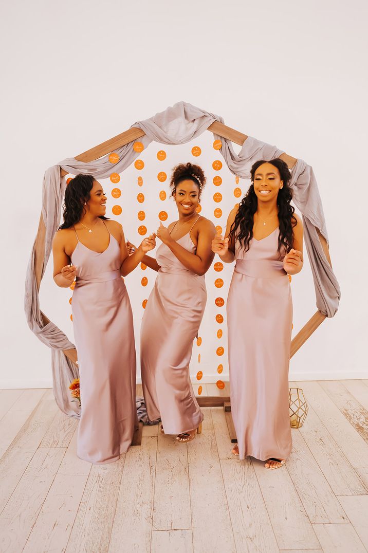 Pink satin bridesmaid dresses worn by bridesmaids standing in front of hexagonal backdrop decoration 