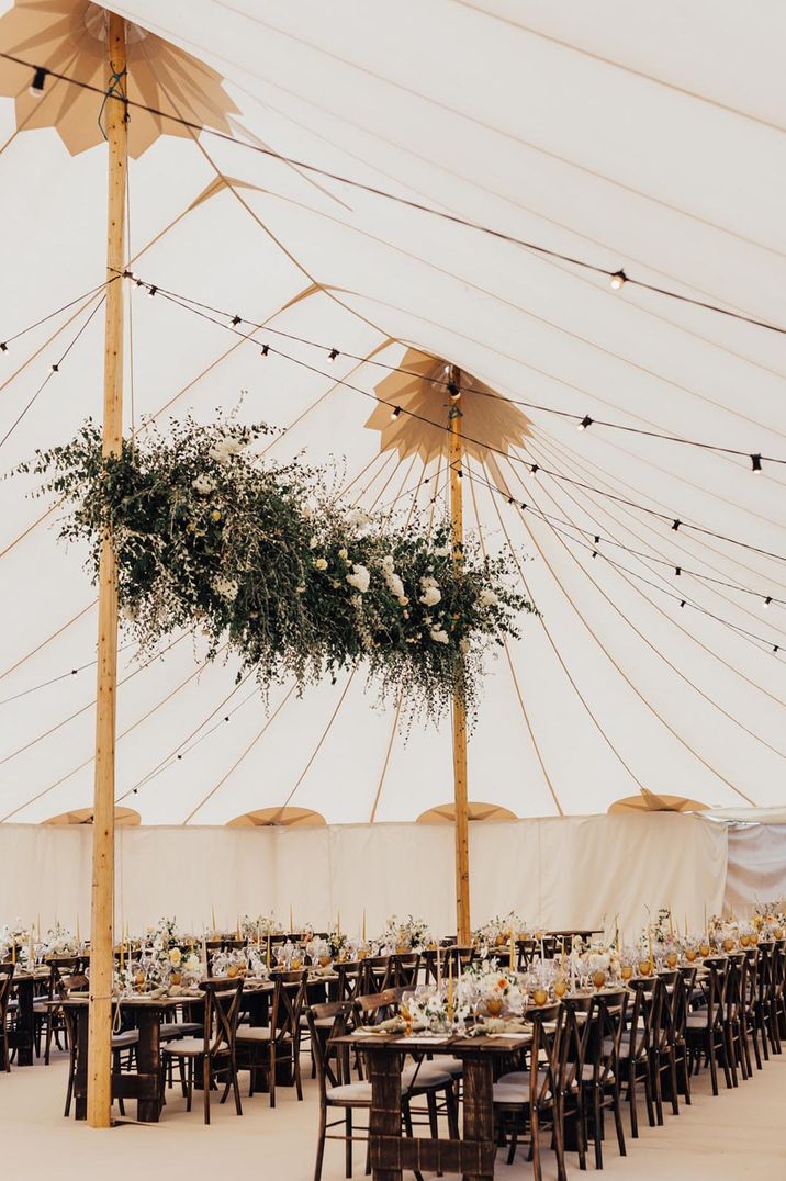 Marquee wedding with festoon lighting and large hanging foliage installation with long banquet tables with yellow wedding table decor 