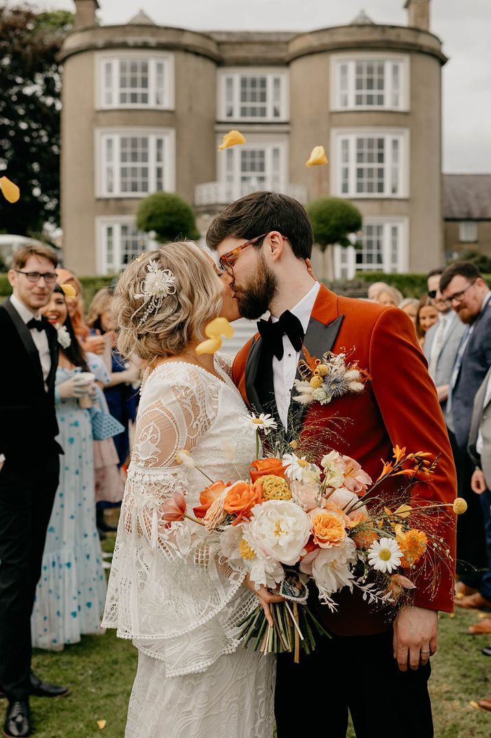 Groom in velvet suit jacket with bride in lace wedding dress carrying orange and yellow wedding bouquet 