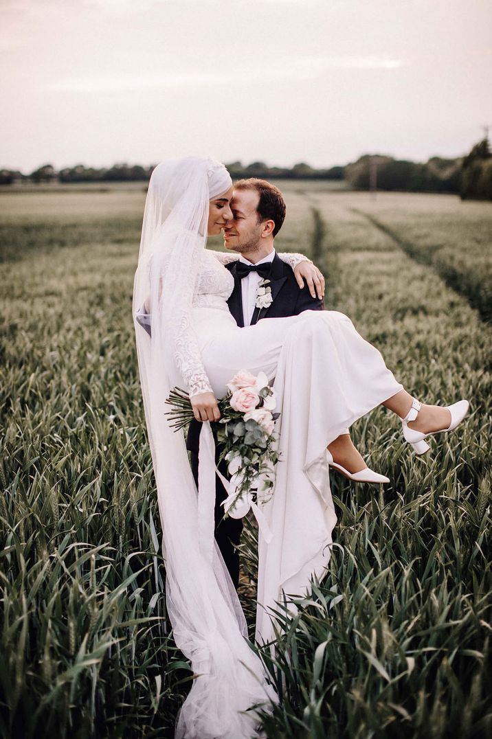 Turkish groom in a tuxedo picking his bride up in a long sleeve lace wedding dress in a field 