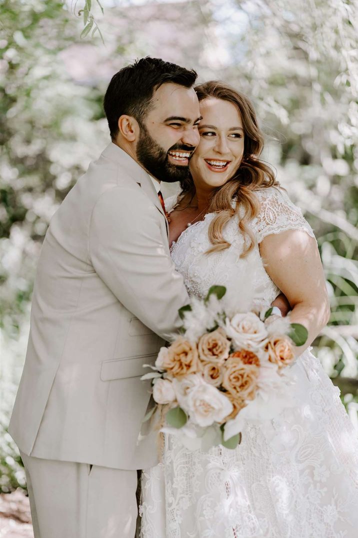 Groom in light grey suit and bride in DIY short sleeved lace wedding dress embracing at garden wedding