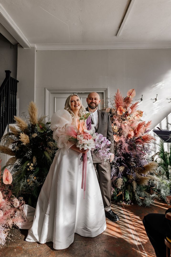 Bride in statement puff sleeve dress and groom in grey suit in front of the dyed pampas grass purple and pink flower columns with anthuriums 