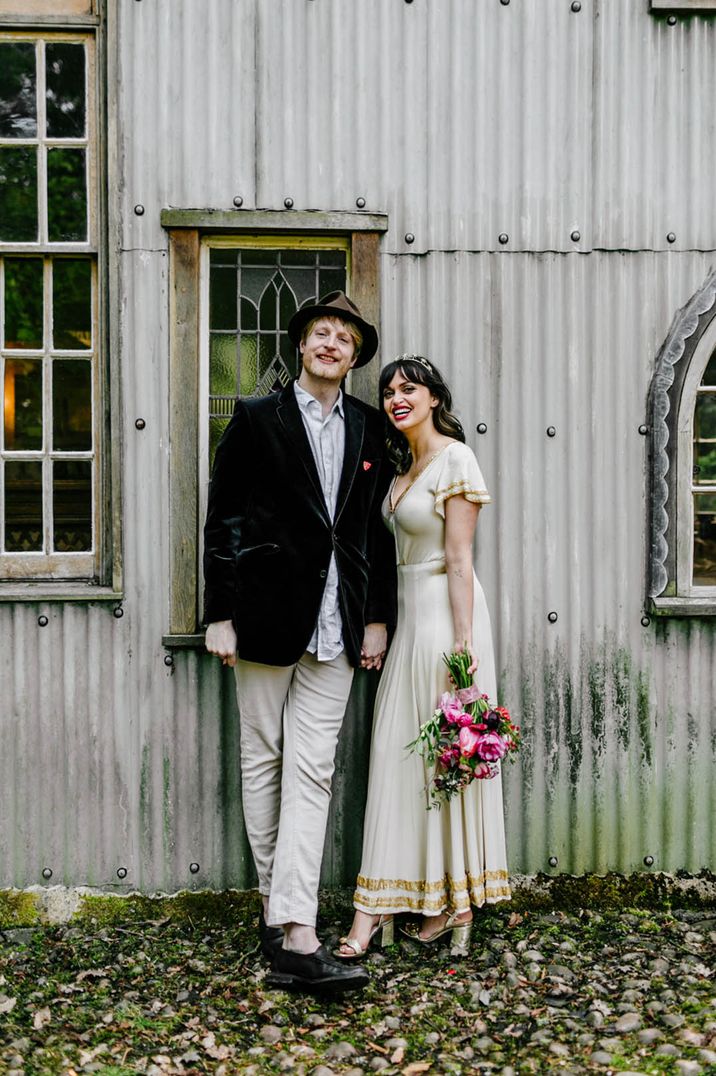 Groom in velvet suit jacket and hat standing with the bride in a vintage wedding dress with gold detailing 