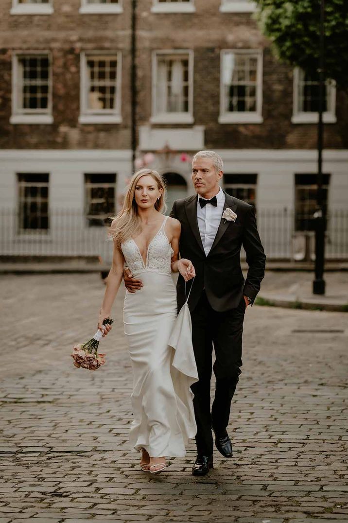 Bride in lace and satin sleeveless wedding dress walking with groom in classic black tux and white boutonniere walking around outside of The Zetter Townhouse - one of the wedding venues London