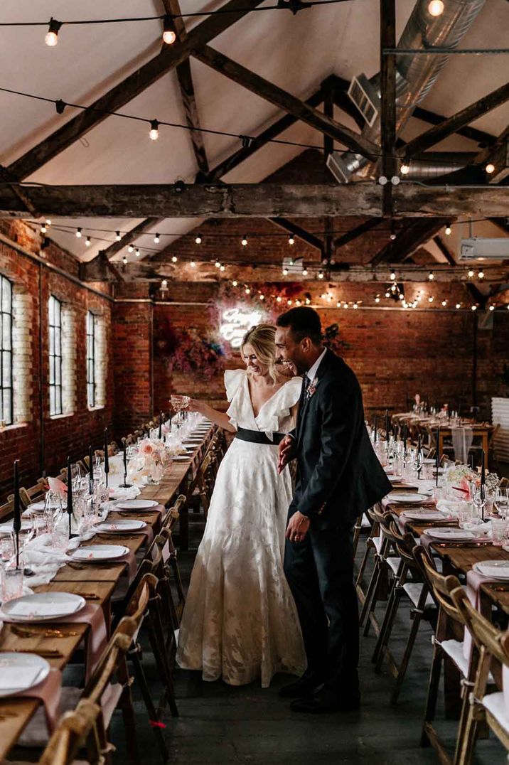 Bride in short sleeve ruffle wedding dress and groom in classic groom tuxedo walking through the neutral minimalistic wedding tablescapes with black tapered candles and pink flower centrepieces 