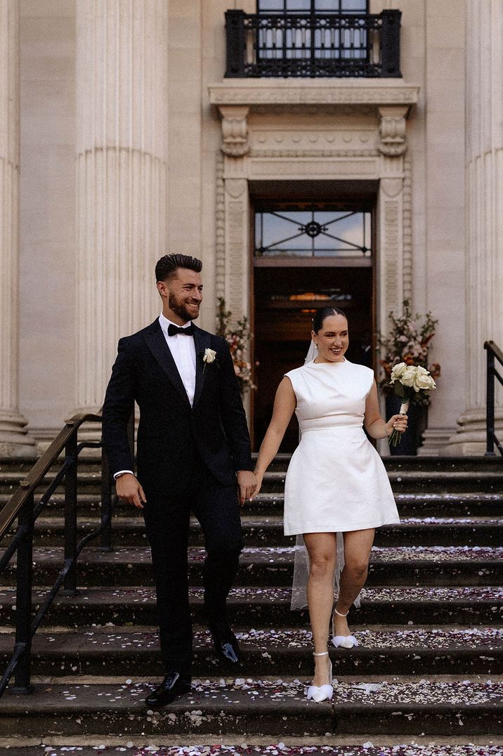 Groom in black tuxedo with white rose buttonhole and bride in short wedding dress and bow wedding shoes coming down the stairs of registry office