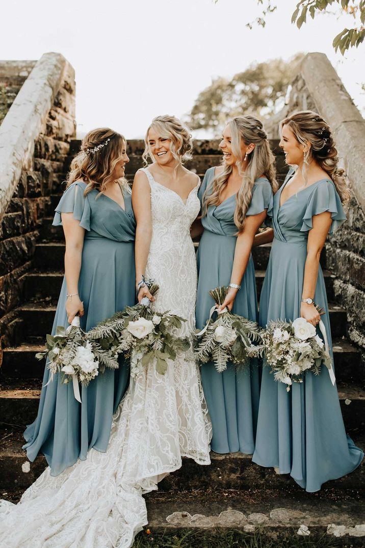 Bride in strappy lace wedding dress standing with bridal party wearing wrap front short sleeve blue bridesmaid dresses 