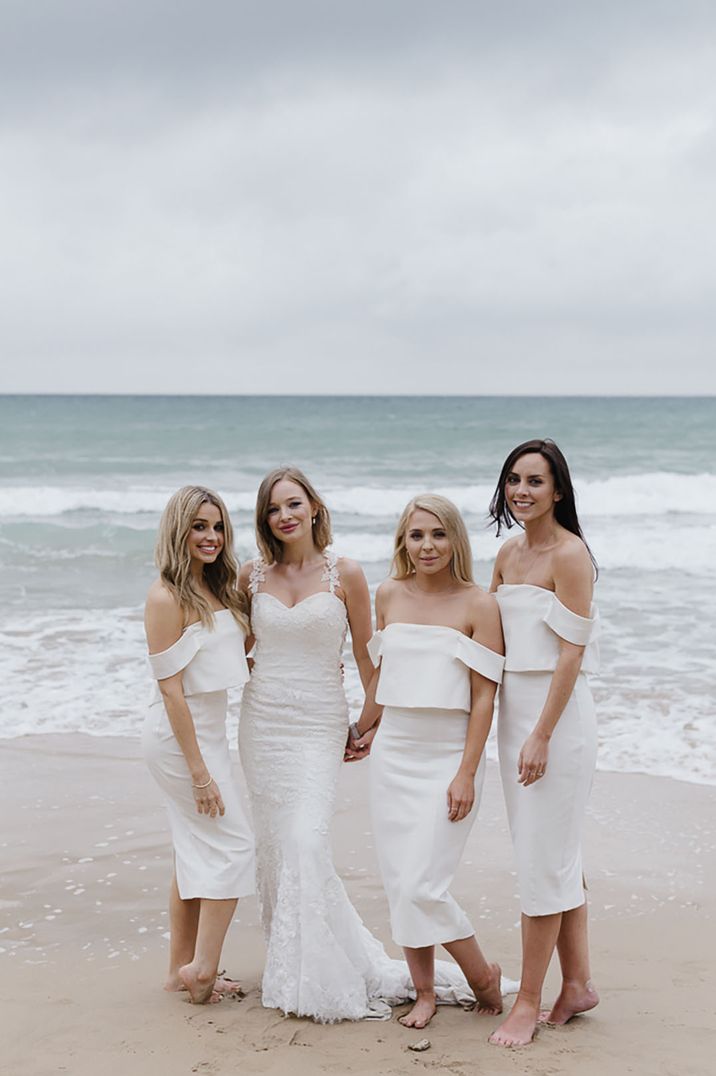 Bridesmaids in white off the shoulder midi bridesmaid dresses posing on the beach with the bride 