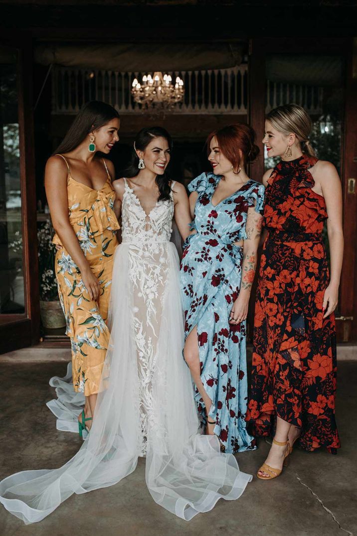 Bride in sleeveless lace and tulle wedding dress standing with bridesmaids in mismatched colourful floral patterned bridesmaid dresses 