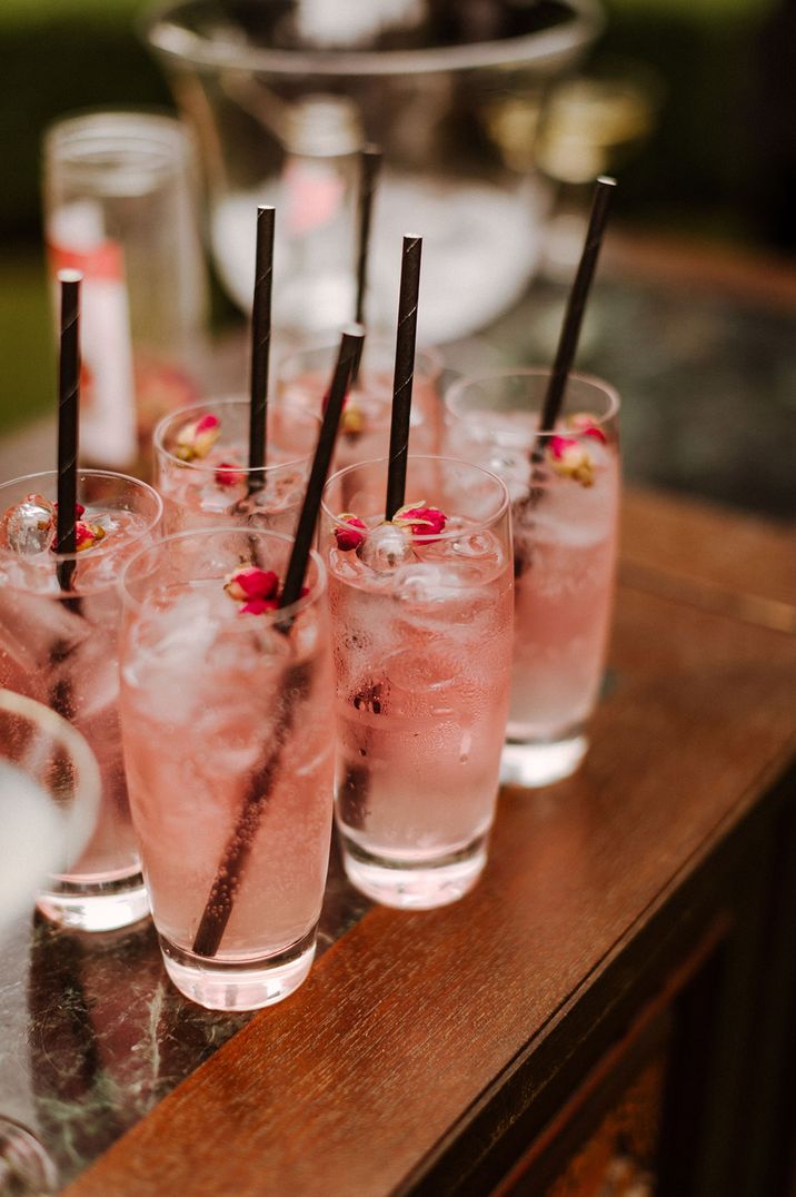 Tall glasses filled with pink cocktails with black straws, ice cubes and edible flowers 