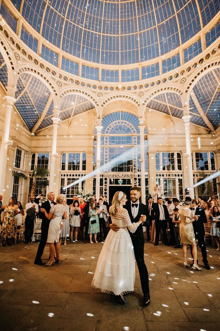 Bride in a lace and polka dot wedding dress dancing in the conservatory at Syon park with her husband in a black tuxedo 