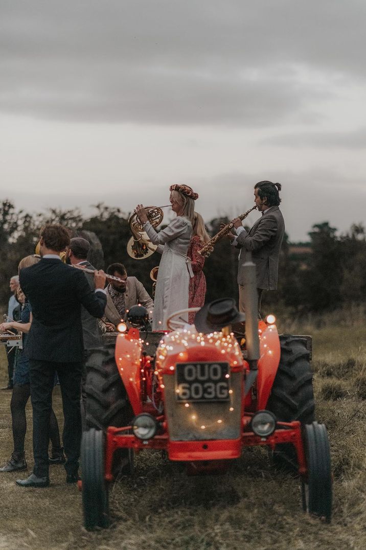 Couple on red tractor with fairy lights, couple playing brass instruments, bride in pink floral crown and long sleeve wedding dress