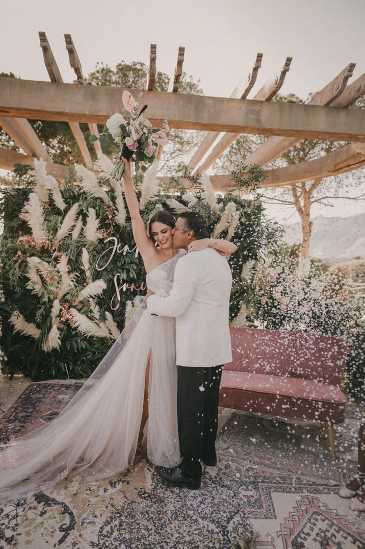 Groom in white tuxedo smiling at the bride in tulle wedding dress lifting bouquet in the air next to pampas grass arrangement and sign 