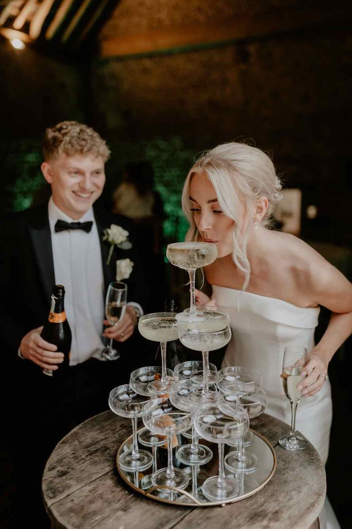 Bride in strapless satin wedding dress sipping from wedding champagne tower with groom in classic black tuxedo 