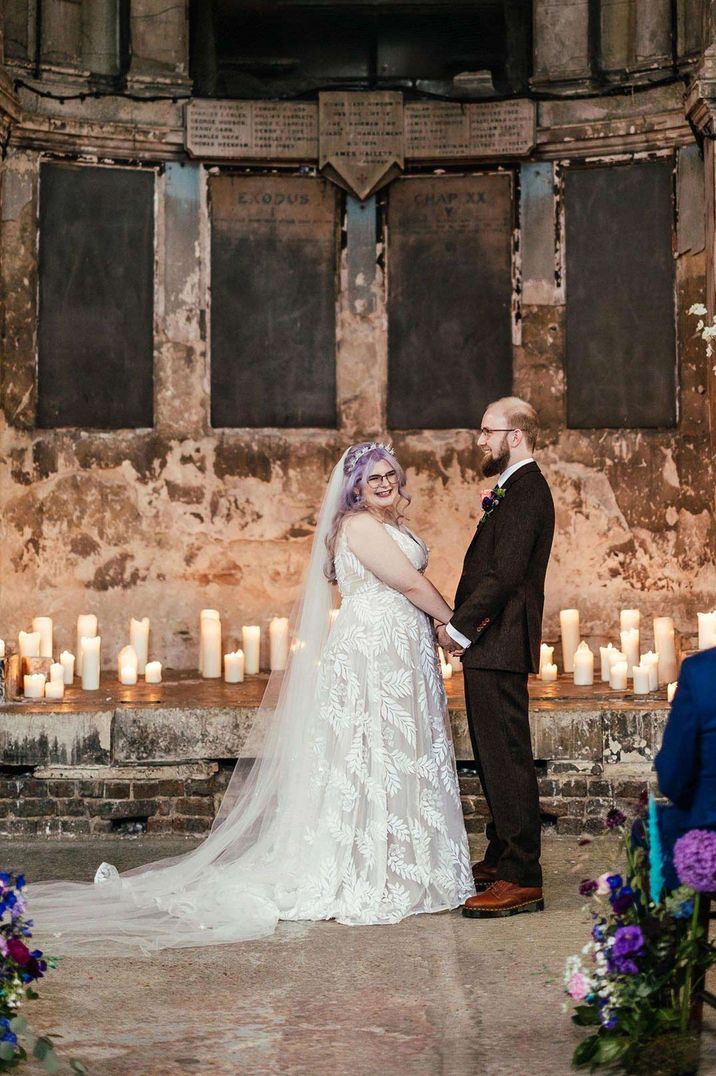 Bride in Evie Young bridal gown and groom in dark brown grooms suit at the alter of alternative UK wedding venue with pillar candles 