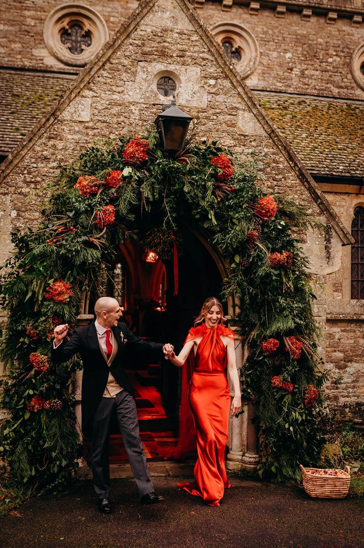 Groom and bride in red satin wedding dress exit from their church ceremony with red flower arch decorating the entrance 