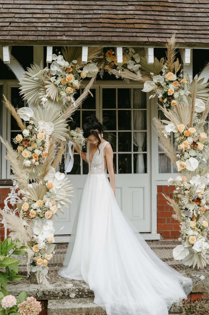 Bride stands under enormous arch of peach and cream roses, lillies and foliage by wedding florist Peony & Thistle Designs