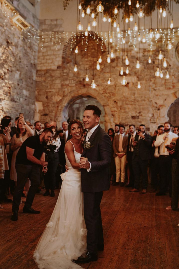 Bride in a satin slip wedding dress with appliqué skirt overlay dancing under a canopy of Edison bulb at their first dance 