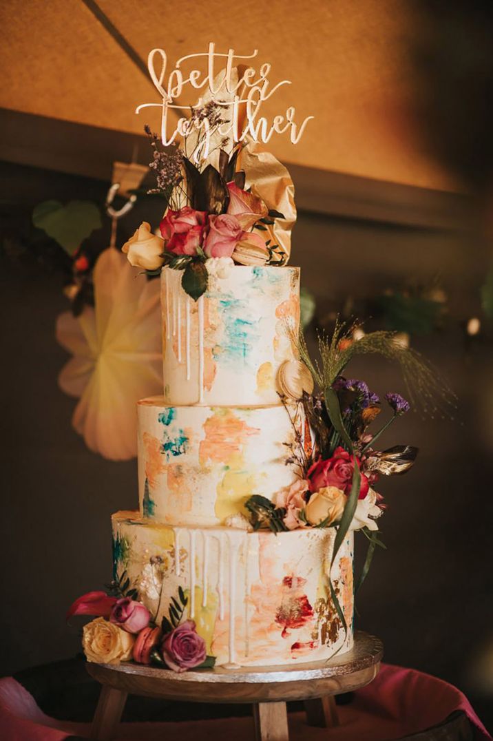 Three tier white wedding cake with colourful paint pattern with blues, pink, peach, gold icing with flowers and gold cake topper