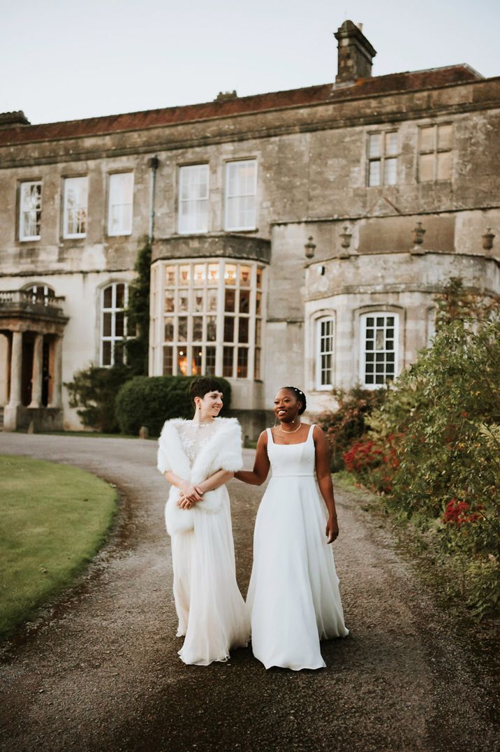 Two brides in complimenting bridal outfits walking the grounds of Elmore Court wedding venue