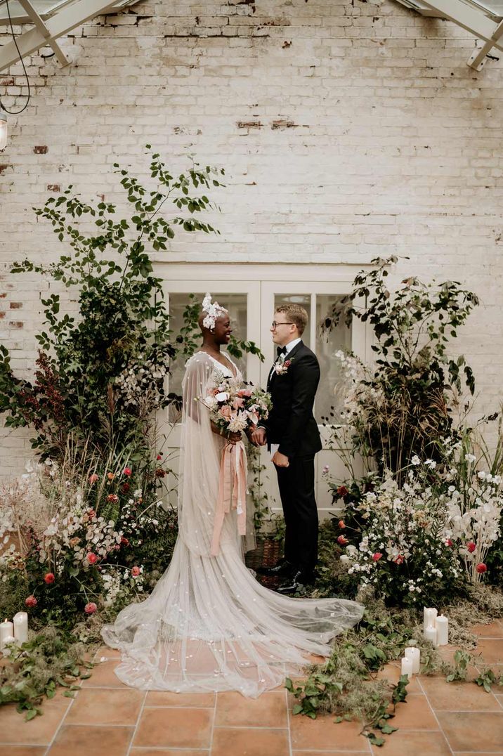 Bride in long sheer wedding dress with sheer cathedral length veil and bridal crown and groom in classic black tuxedo standing in glasshouse wedding venue - book a wedding venue 