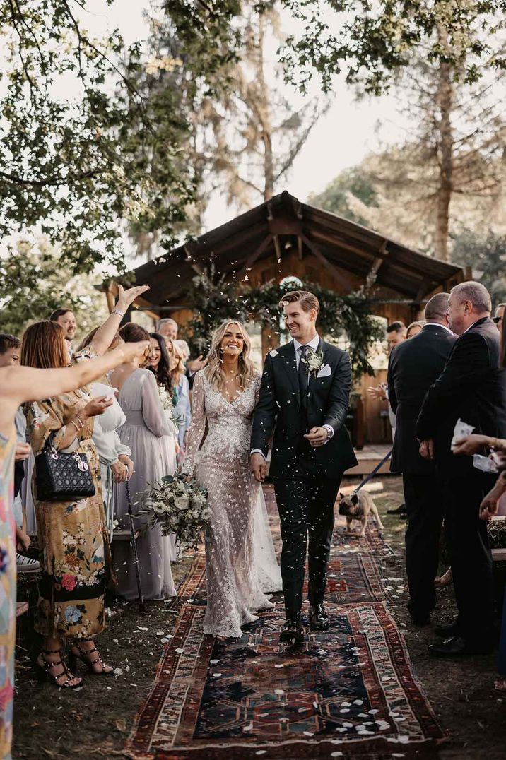 Bride in boho long sleeve sheer embellished wedding dress holding rose, peony, dried flower and eucalyptus bridal bouquet walking down the aisle doing petal confetti throw with groom in three piece suit at Oak Tree Barn wedding venue 