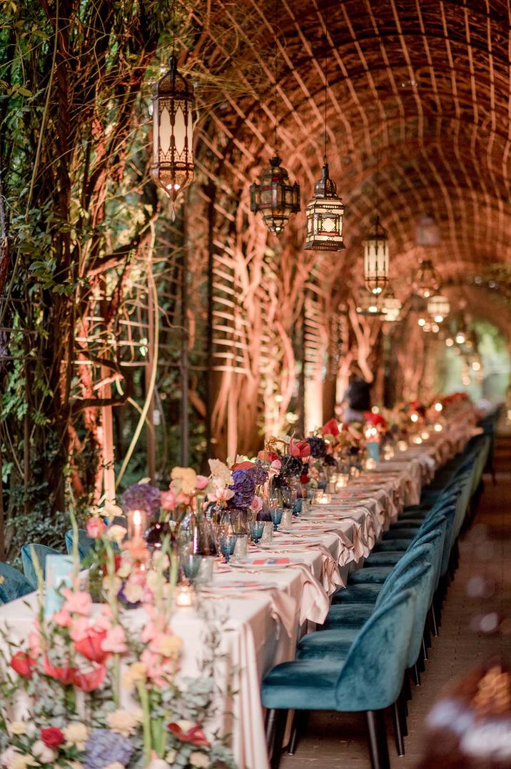 Long banquet table with blue velvet wedding chairs decorated with colourful waterfall wedding flowers with blue coloured glassware 