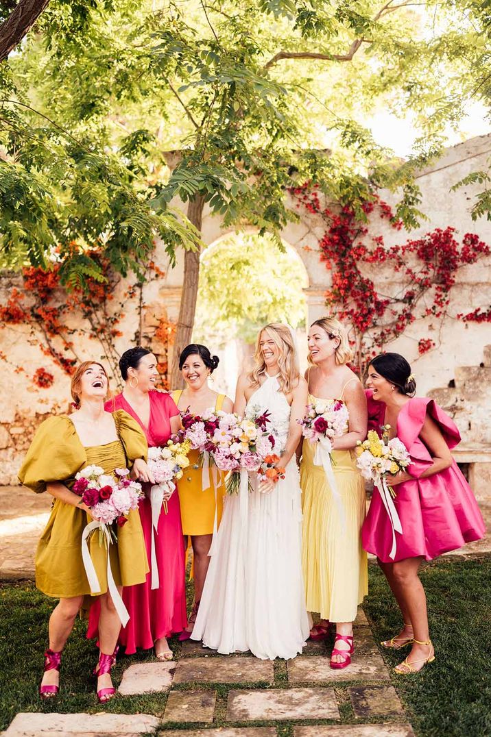 Bride wearing halterneck wedding dress standing with bridesmaids in vibrant mismatched bridesmaid dresses - hen party ideas