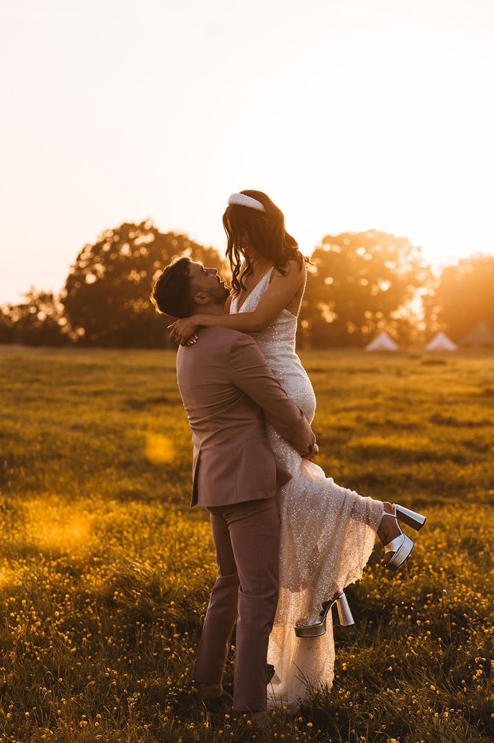 The bride in a sparkly wedding dress and metallic silver platform wedding shoes being picked up by the groom for sunset wedding photo