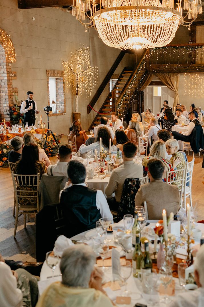 Wedding guests seated for the wedding breakfast with the groom reading out his wedding speech at NI wedding venue with chandelier 