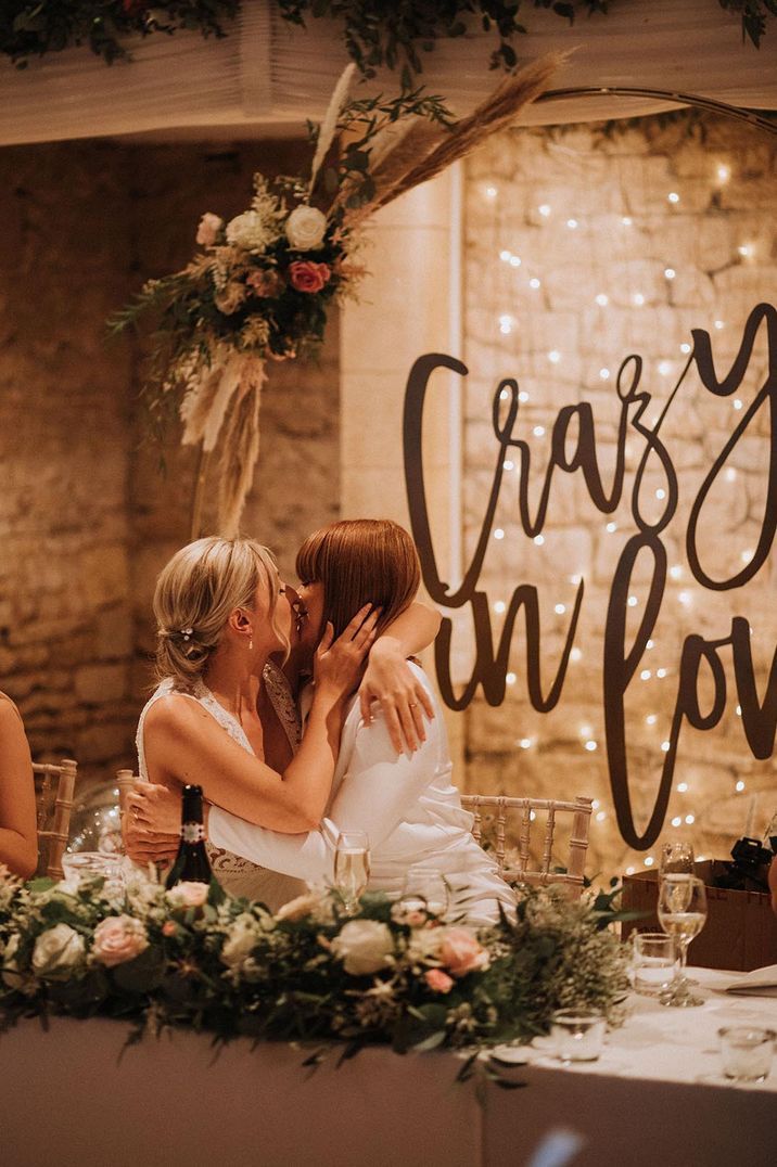 two brides kissing during the speeches at their rustic barn wedding with a giant Crazy In Love sign behind the top table 