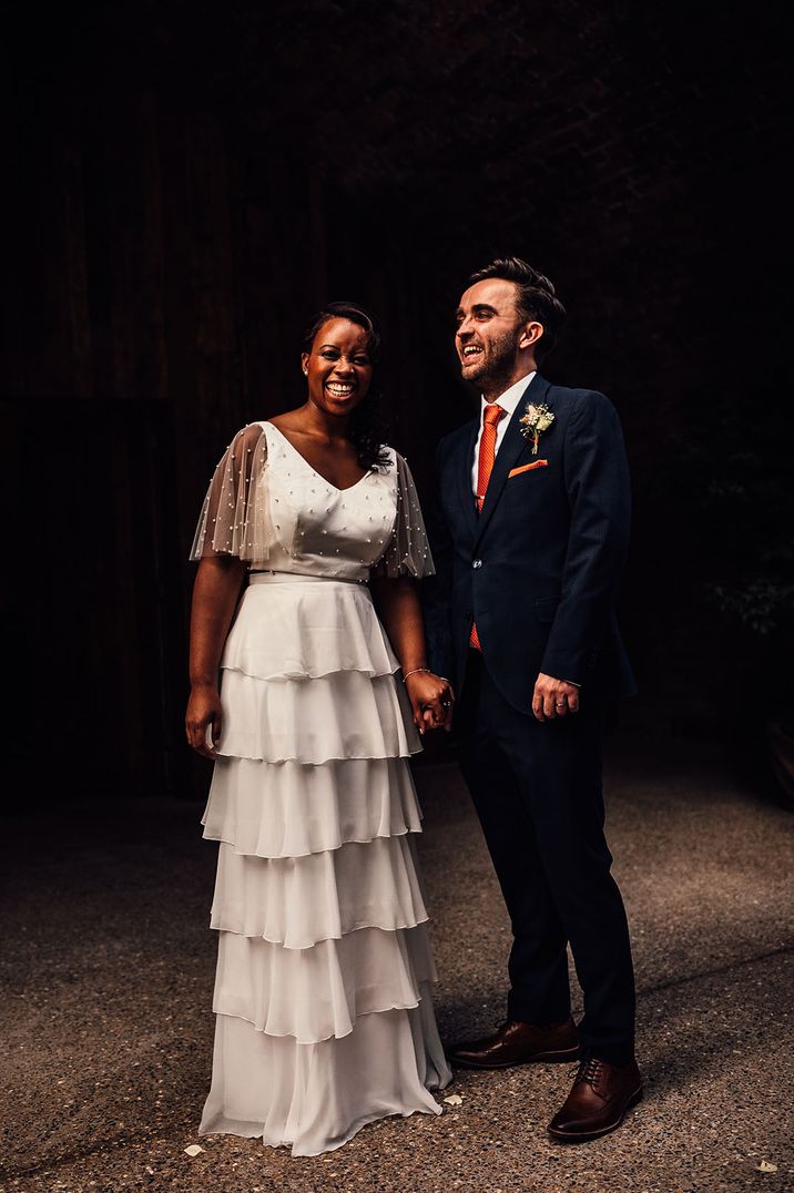 Bride in ruffle skirt and pearl top with groom in blue suit and orange tie holding hands