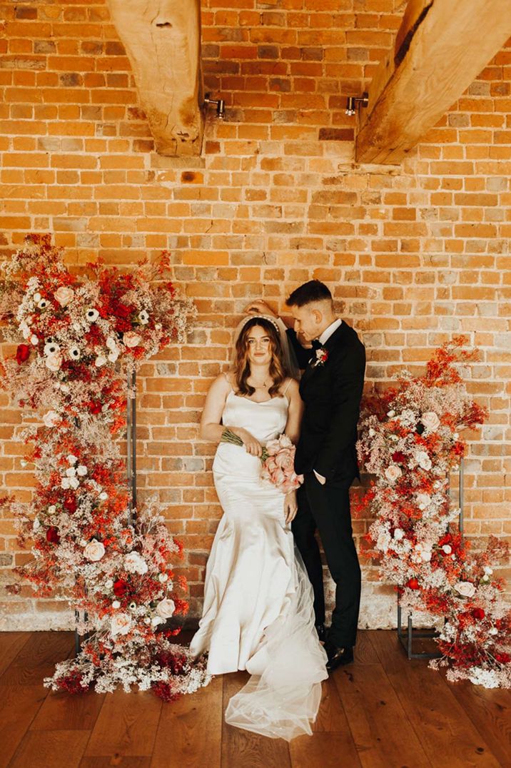 Bride in satin look wedding dress and pearl headband and groom with black tux standing by orange, red and white floral arch