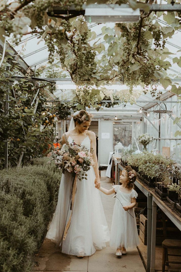 Bride in sleeveless satin wedding dress holding large dried flower neutral bouquet and flower girl walking through The Secret Herb Garden Edinburgh glasshouse wedding venue 