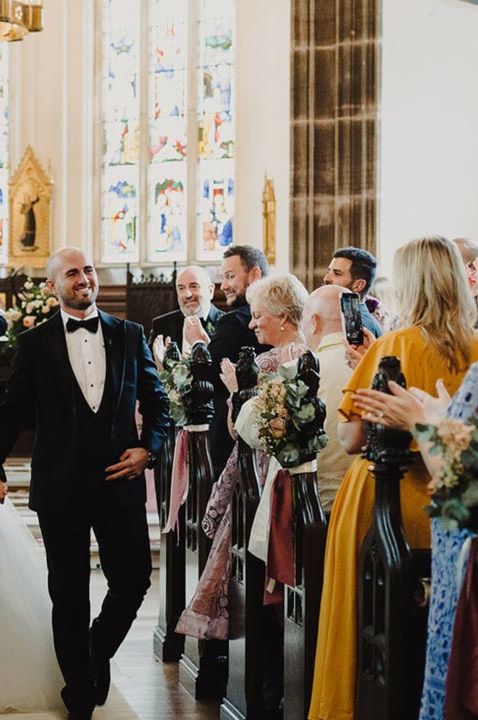 Bride in v neck sleeveless wedding dress with tulle skirt walking down the aisle with groom in classic black tuxedo at Cluny Castle wedding venue 