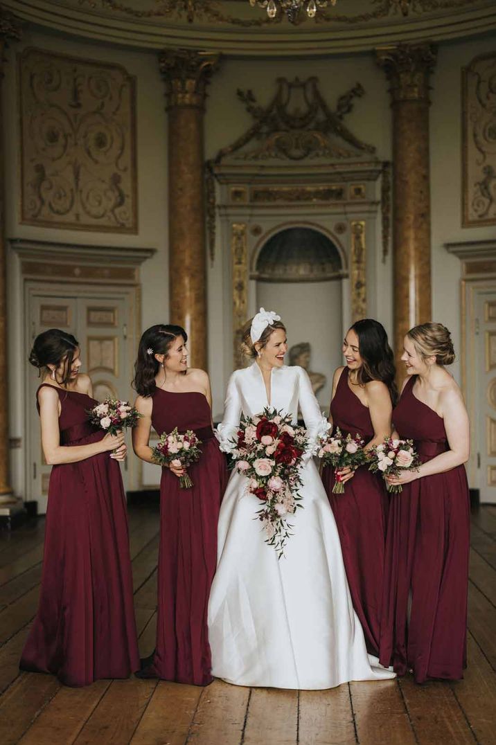 Bride in classical long sleeve satin wedding dress standing with bridesmaid in one shoulder burgundy bridesmaid dresses at Stowe House wedding