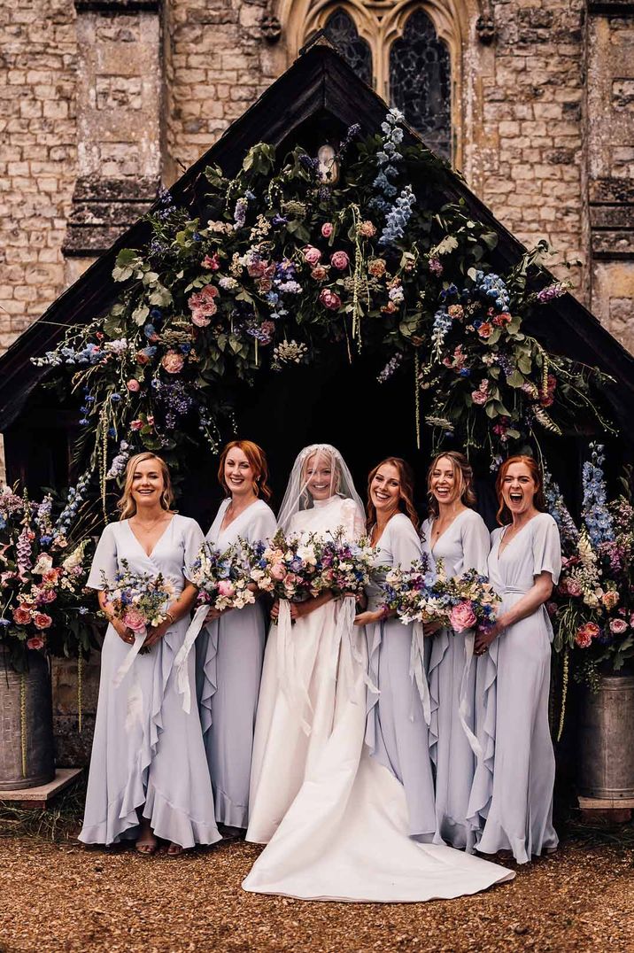 Bride in long sleeve wedding dress standing outside church with bridesmaids in short sleeve lilac front wrap bridesmaid dresses