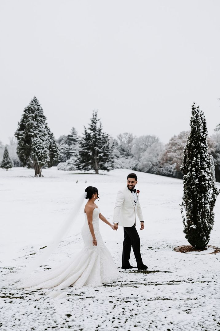 Bride in mermaid wedding dress walking with groom in white tuxedo at snowy wedding 