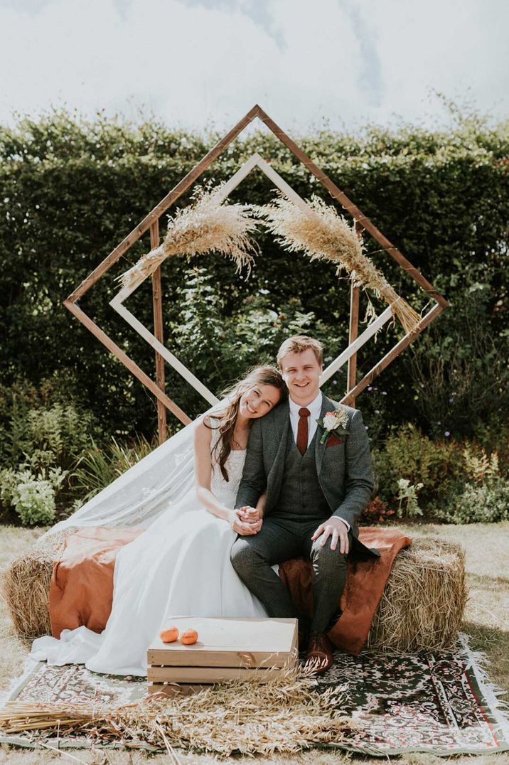 Bride in Bardot top wedding dress and veil and groom in dark suit sitting by DIY geometric wedding decoration