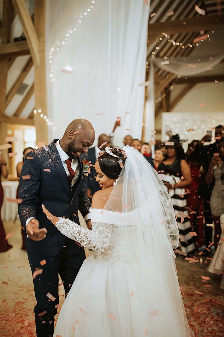 Bride in off shoulder lace wedding dress and groom in dark suit and red tie dancing with confetti