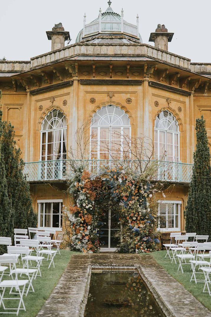 Exterior of Sezincote house glasshouse wedding venue with long, rectangular pond, yellow building and green tinted balcony with large autumnal floral arch 