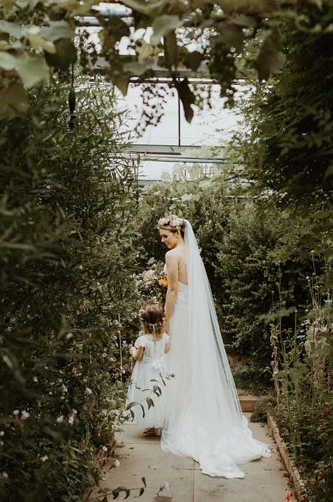 Bride in church length veil walking through glasshouse wedding venue with flower girl 